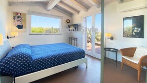 Un dormitorio con cama, una silla, una mesita y un ventanal.