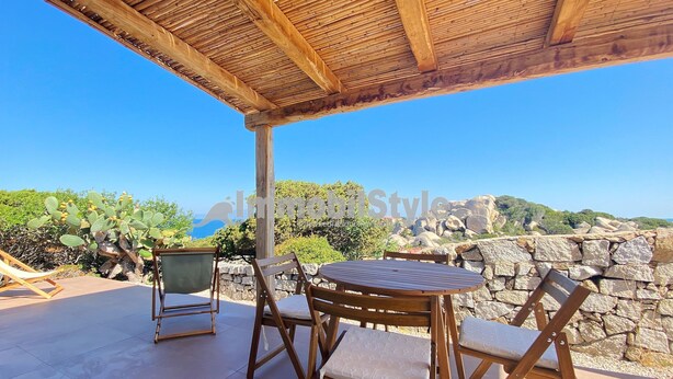 Una mesa y sillas de madera bajo una pérgola, con un muro de piedra y follaje al fondo.