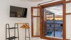 A room with a TV mounted on the wall, a window with a view of a building and a pool, and a metal shelf with hangers. - La Si'Ma, with private pool, 2-bedrooms close to Margarites Village (Plevriana)