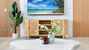 Ein modernes Wohnzimmer mit einem großen Flachbildfernseher an einer weißen Wand, einer Holzkabine mit offenen Regalen, einer weißen Vase mit Blumen und einer Topfpflanze.