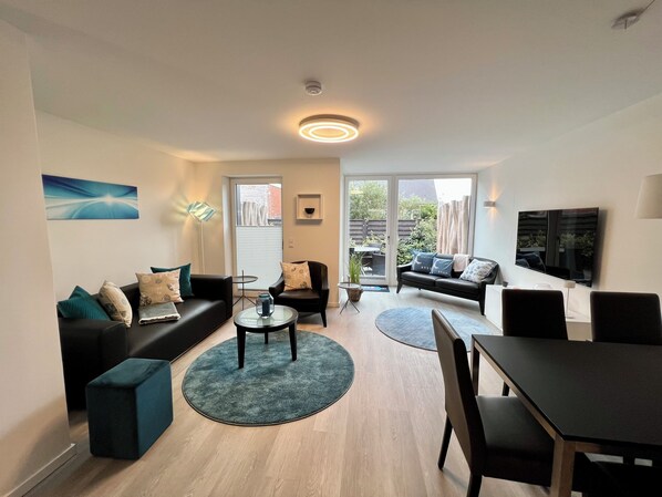 A modern living room with a sofa, chairs, a coffee table, and a TV. There is a sliding door leading to a balcony with outdoor furniture.