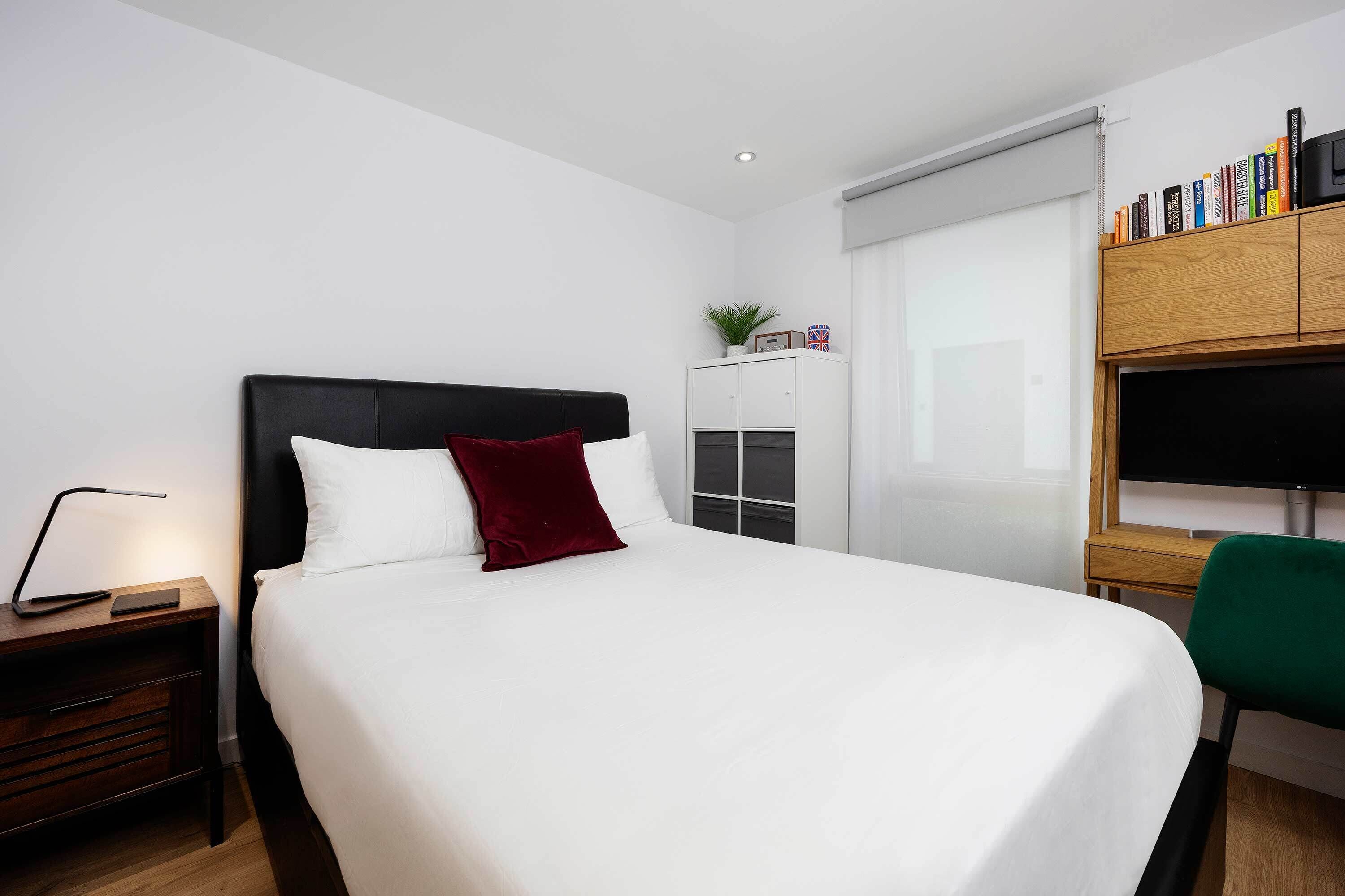 Een keurig opgeruimde hotelkamer met een groot bed, een nachtkastje met een lamp, een klein kastje met boeken en een groene stoel.