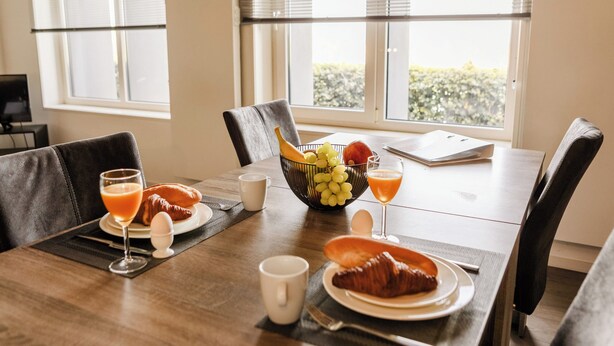 Un desayuno con una mesa de madera, sillas, un bol de fruta, croissants y zumo de naranja.