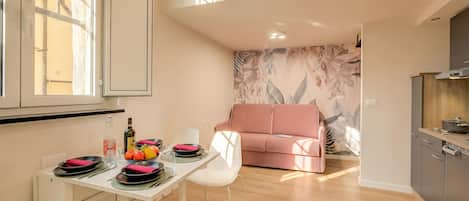 A dining area with a table set for two, a pink sofa, and a kitchen area with a stove.