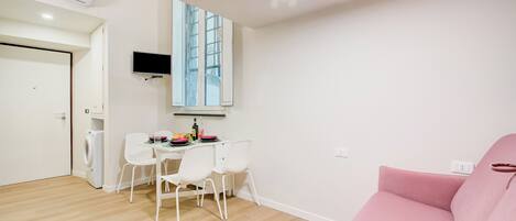 Un salon moderne avec un canapé rose, un coin repas avec une table et des chaises, un lave-linge et un téléviseur fixé au mur.