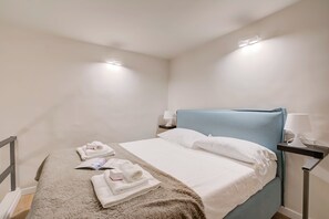 A neatly made bed with white linens and a blue headboard, a bedside table with a lamp, and a small nightstand with a lamp. - Old Town Maison - City Center (Genoa)