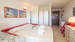 Habitació d'hotel amb un llit, escriptori i cadira. Hi ha dos quadres a la paret, un armari i una porta que condueix a una altra habitació.
