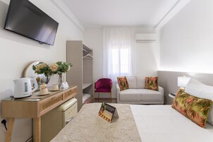 Quarto de hotel com cama, escrivaninha com chaleira, cadeira, sofá, televisão e janela com cortinas.