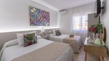 A hotel room with two beds, a desk with a vase of flowers, a painting on the wall, and a television mounted on the wall.