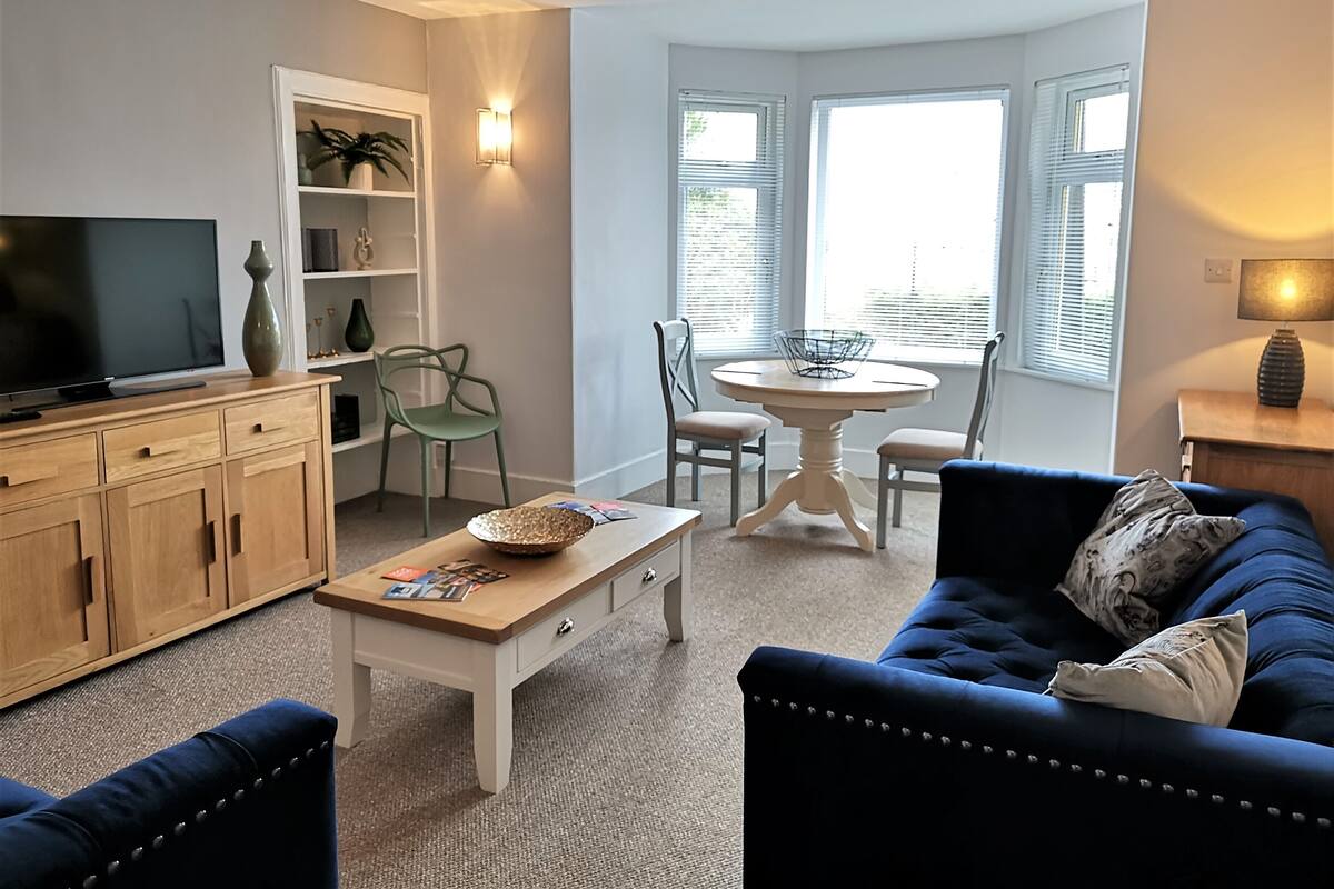 A living room with a TV, wooden cabinets, a blue sofa, a coffee table, and a dining area with a table and chairs.