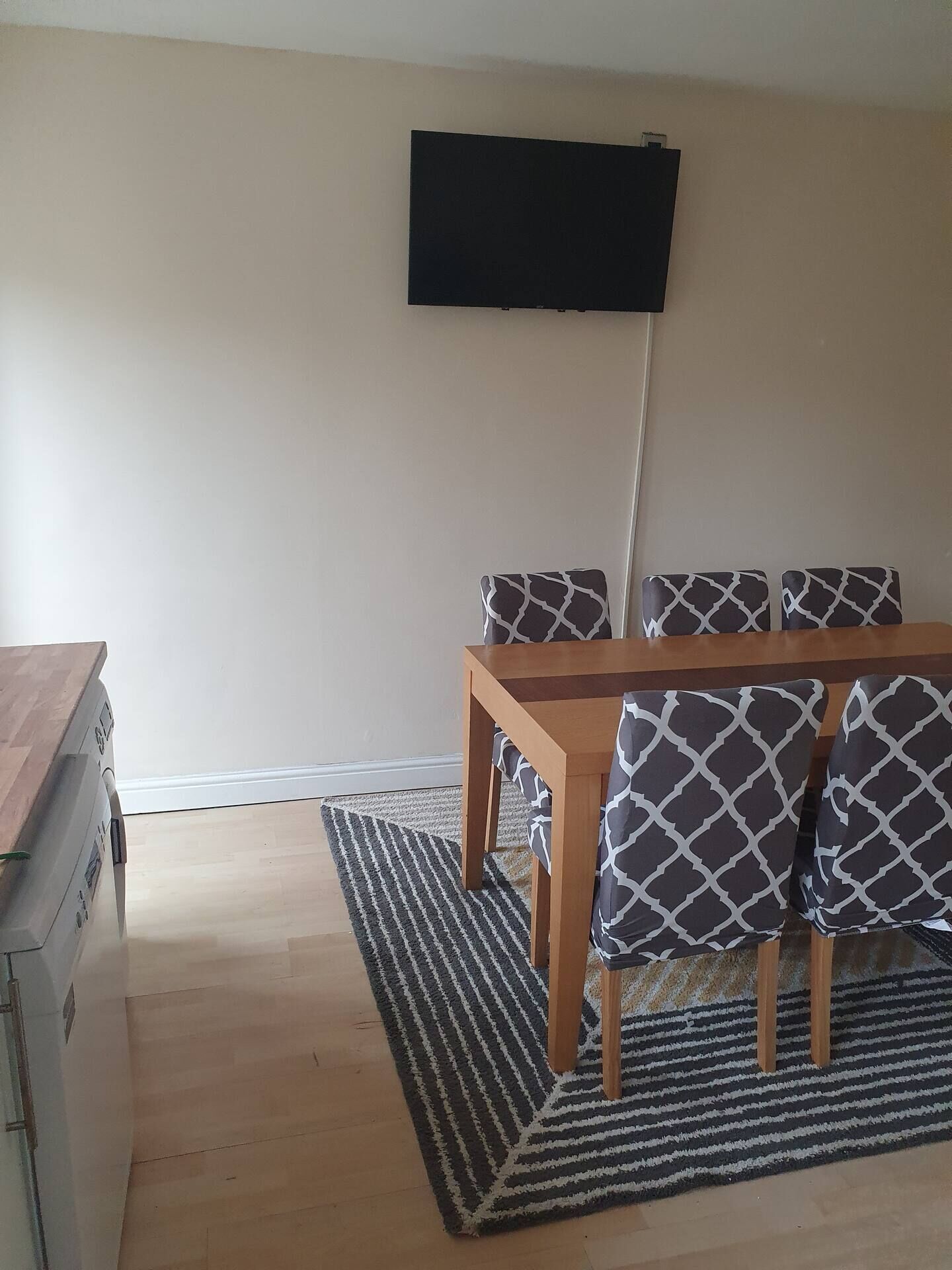 A room with a wooden table and patterned chairs, a flat-screen TV mounted on the wall, and a striped rug on the floor.