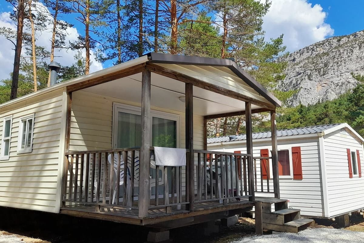 A mobile home with a porch and a small attached cabin, surrounded by trees and a mountain.