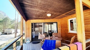 Una cabaña de madera con un porche, un comedor con mesa y sillas, y vista al bosque circundante.