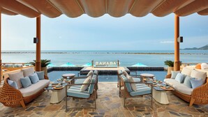 A covered outdoor seating area with a pool, lounge chairs, and a view of the sea.
