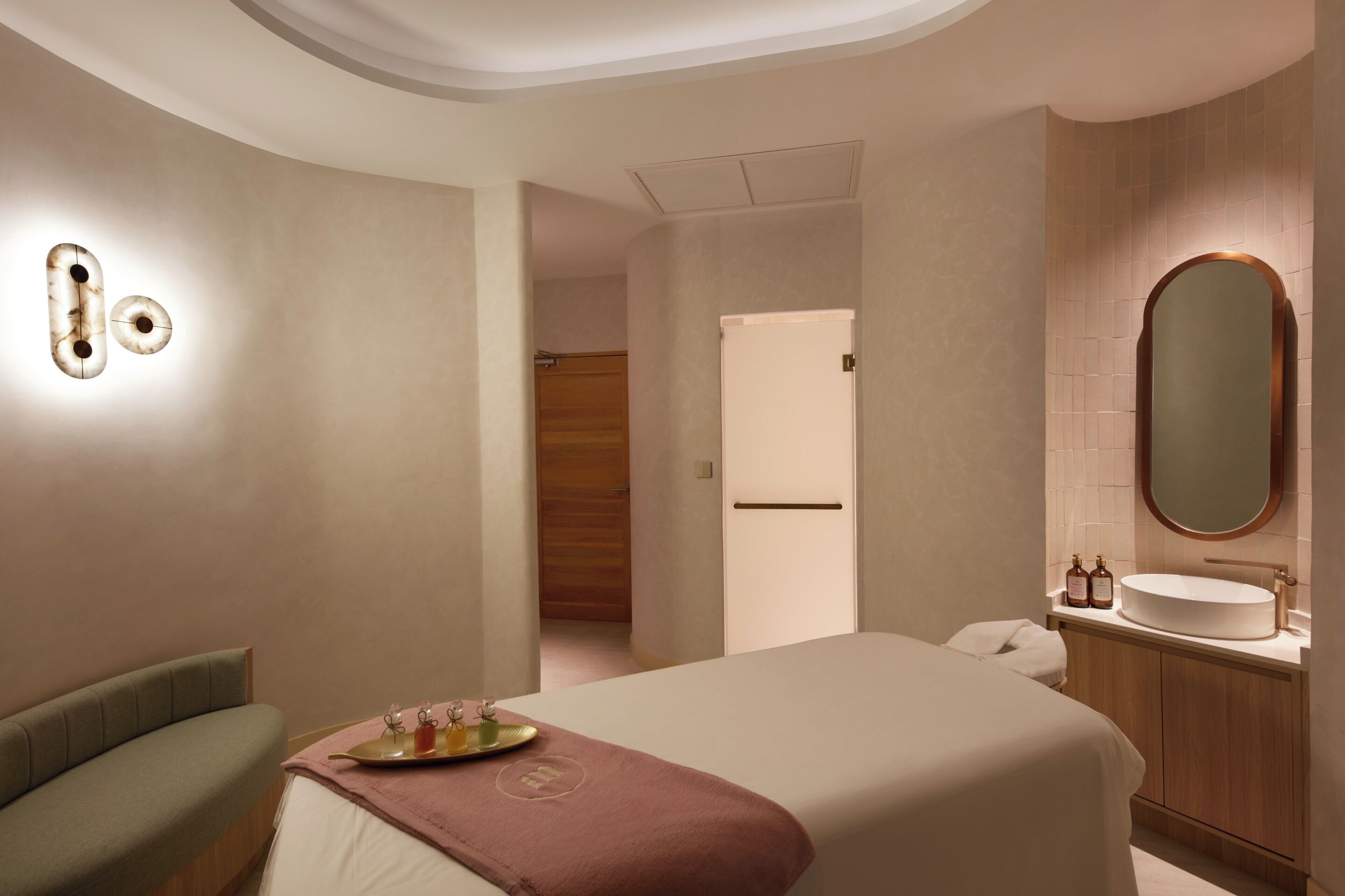 A modern hotel room with a large bed, a bedside table with a tray of toiletries, a wooden bench, a bathroom with a round mirror, and a wooden door.