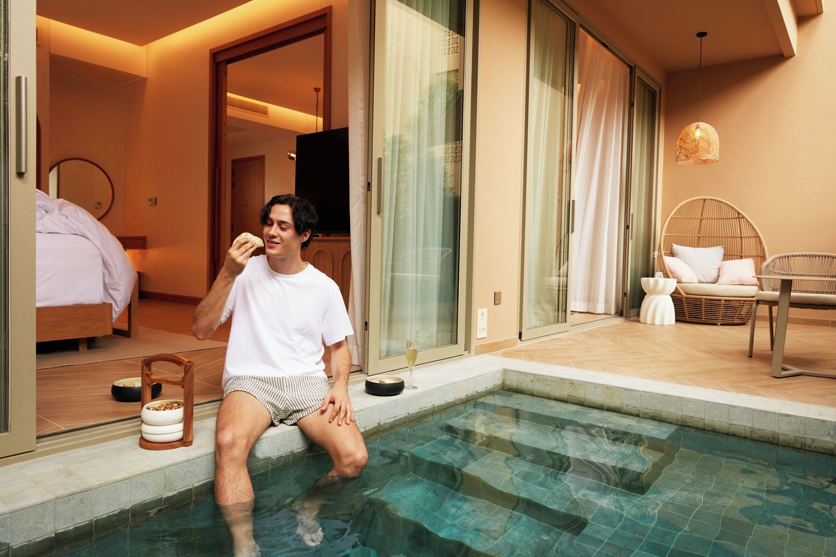 A man sitting by a swimming pool in a modern hotel room with a bed, a dining area, and a wicker chair.