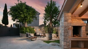 A stone patio with outdoor furniture, a stone fireplace, and a large tree.