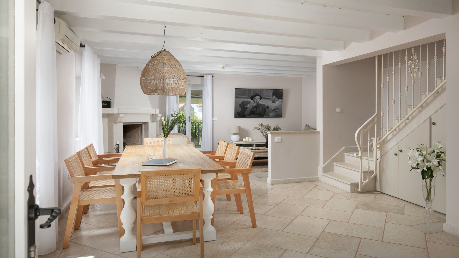 A spacious dining area with a long wooden table and wicker chairs, a hanging light fixture, and a staircase with white railings.