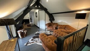Ett rum i loftstil med ett mörkt trägolv, en stor brun lädersoffa, ett glasbord och en lampa. En trappa med ett svart räcke leder upp till en övre våning.