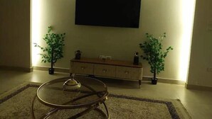 Ett vardagsrum med en platt-TV som är monterad på väggen, ett träskåp, två krukväxter och ett soffbord i glas.