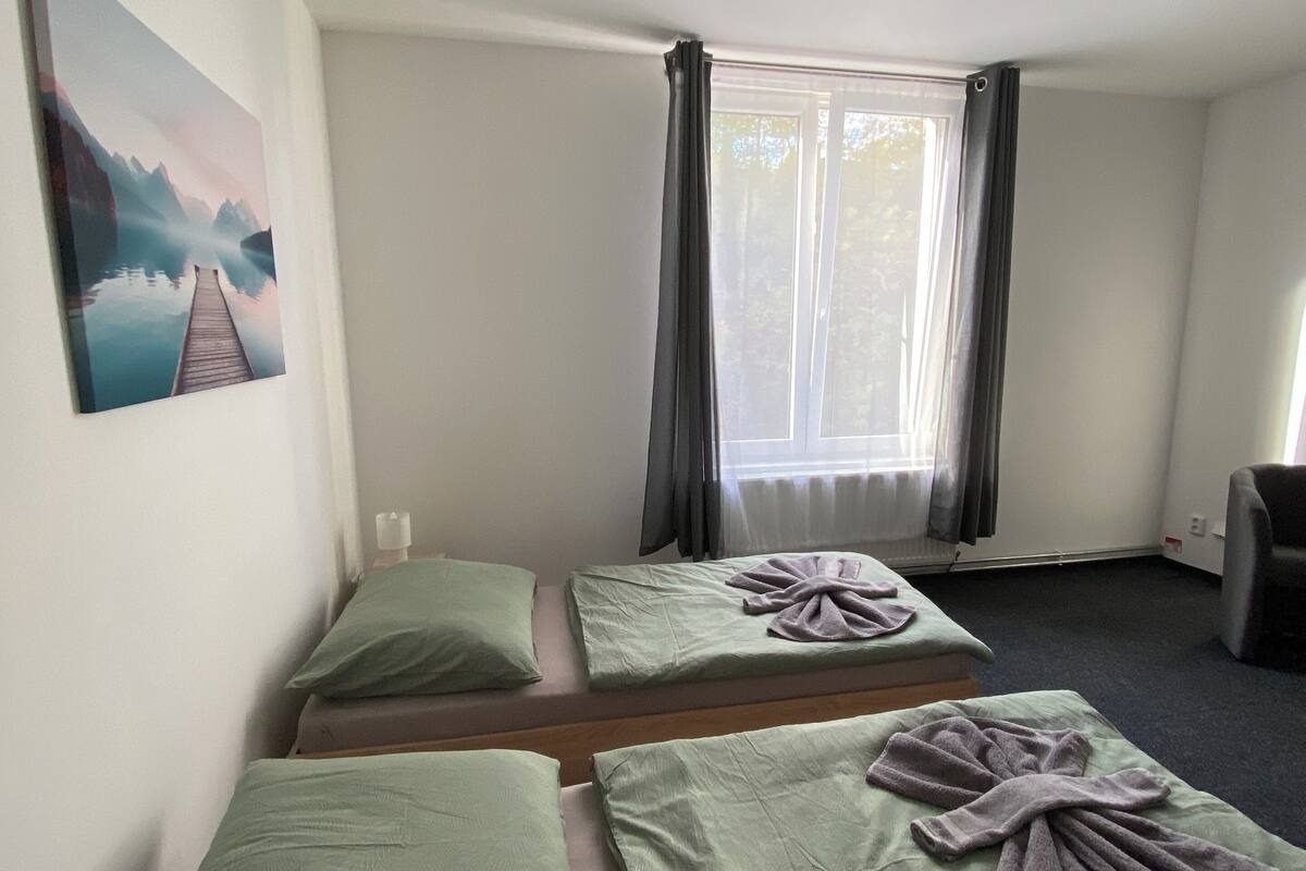 A room with two single beds, a painting on the wall, a window with curtains, and a chair.