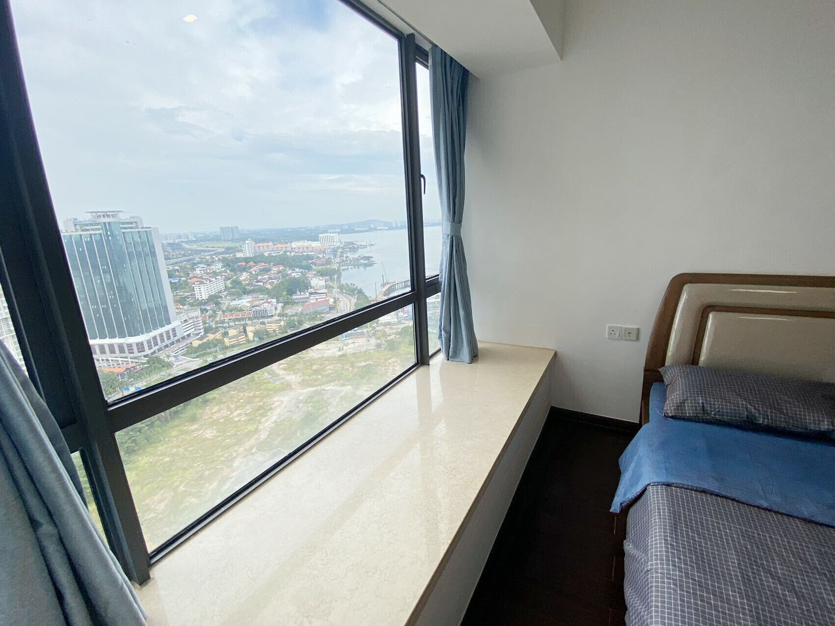 A hotel room with a large window offering a city view, a bed with a headboard, and a bedside table.