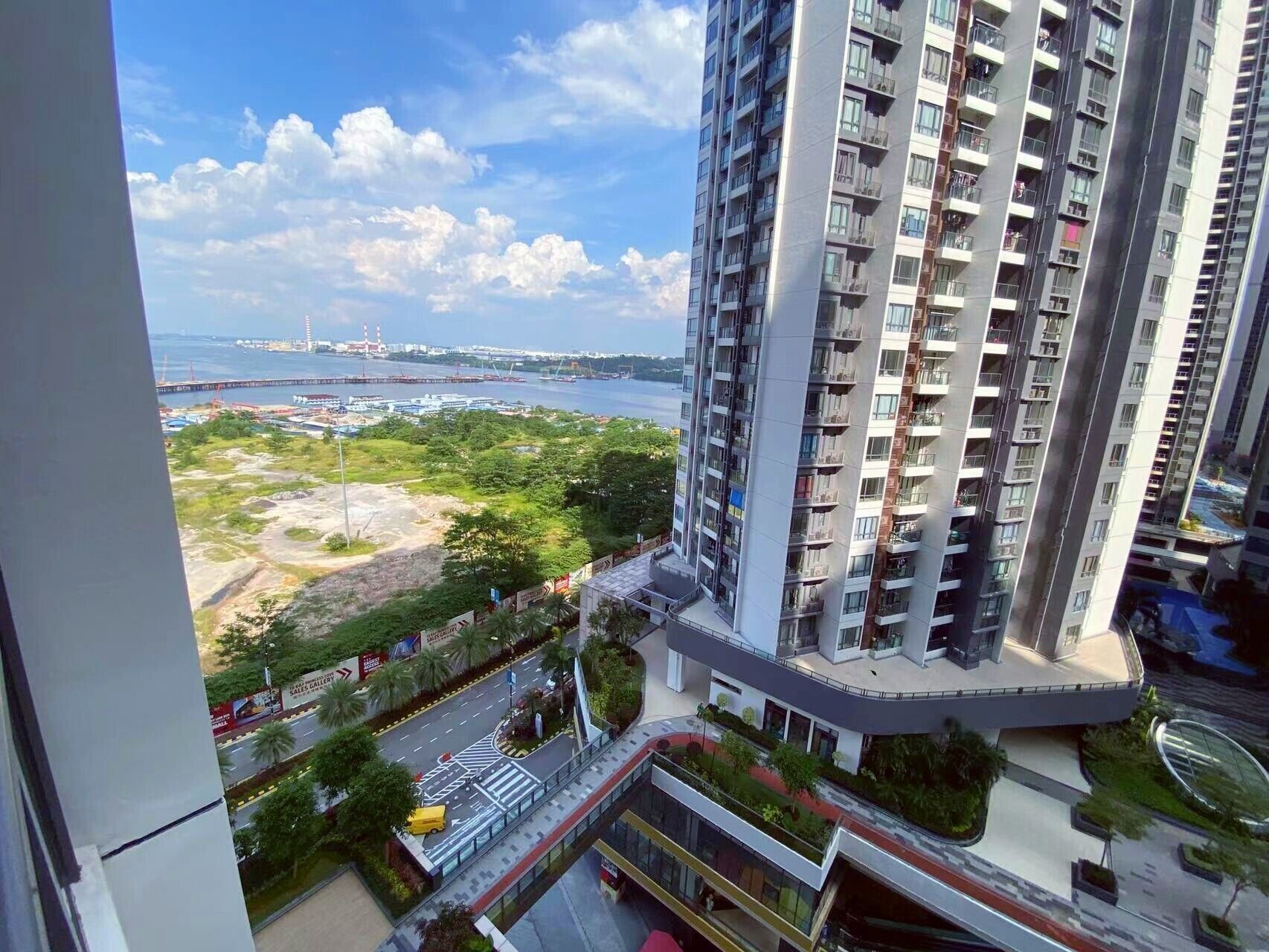 Un immeuble de grande hauteur doté de nombreux balcons, offrant une vue sur un port avec des navires, et comprenant un espace paysager avec des arbres et une route.