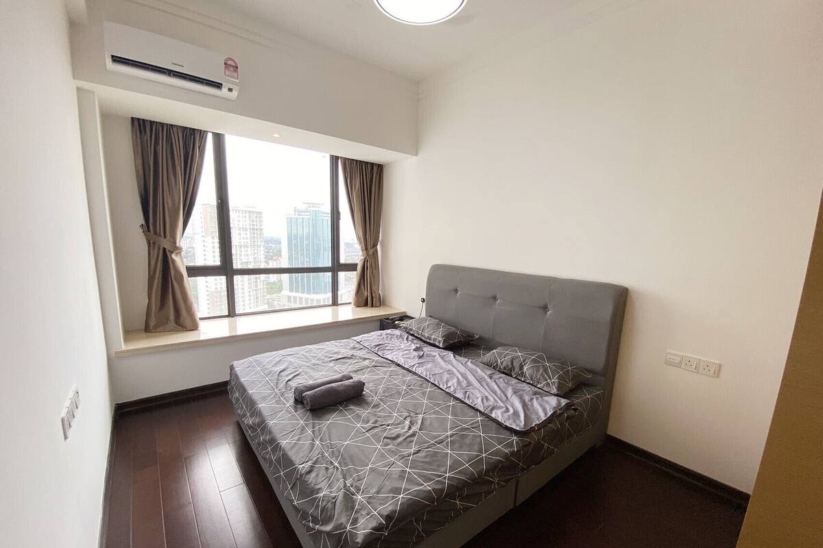 Une chambre à coucher avec un lit, une fenêtre avec des rideaux, un appareil de climatisation et une vue sur des immeubles.