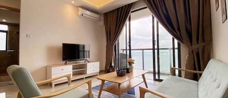 A modern living room with a flat-screen TV, wooden furniture, and a view of the sea through large windows.