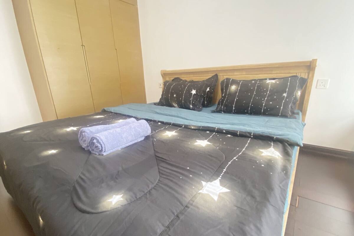 Un lit bien fait, avec une couette foncée et un plaid bleu, une serviette enroulée et des coussins en forme d’étoile.