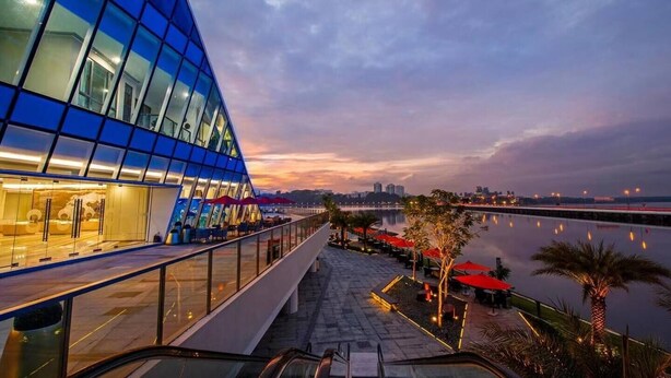 A modern building with a glass facade, a waterfront promenade with outdoor seating, and a view of the city skyline at dusk. - R&F TrexHomestay (Johor Bahru)