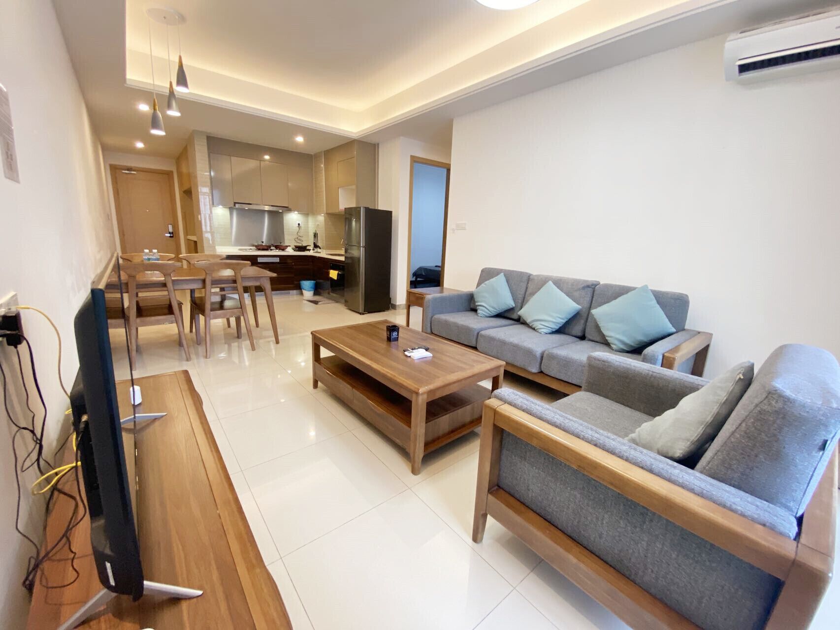 A modern living room with a sofa, coffee table, and TV. There is a kitchen in the background with a refrigerator and a dining area.