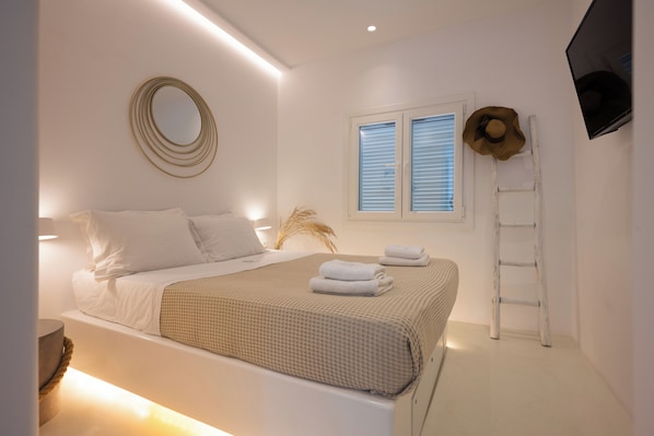 A modern bedroom with a neatly made bed, a wall-mounted TV, and a window with blinds.