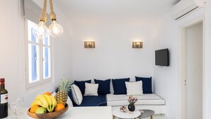 A modern living room with a white sofa, a wooden bowl of fruit, a small round table, and a mounted TV. - Hyades - Downtown Suites No 1 , Mykonos Town (Mykonos Town Cyclades)