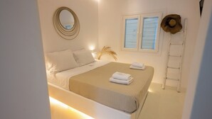 A bedroom with a bed, bedside table, lamp, and a window with blinds. - Hyades - Downtown Suites No 1 , Mykonos Town (Mykonos Town Cyclades)