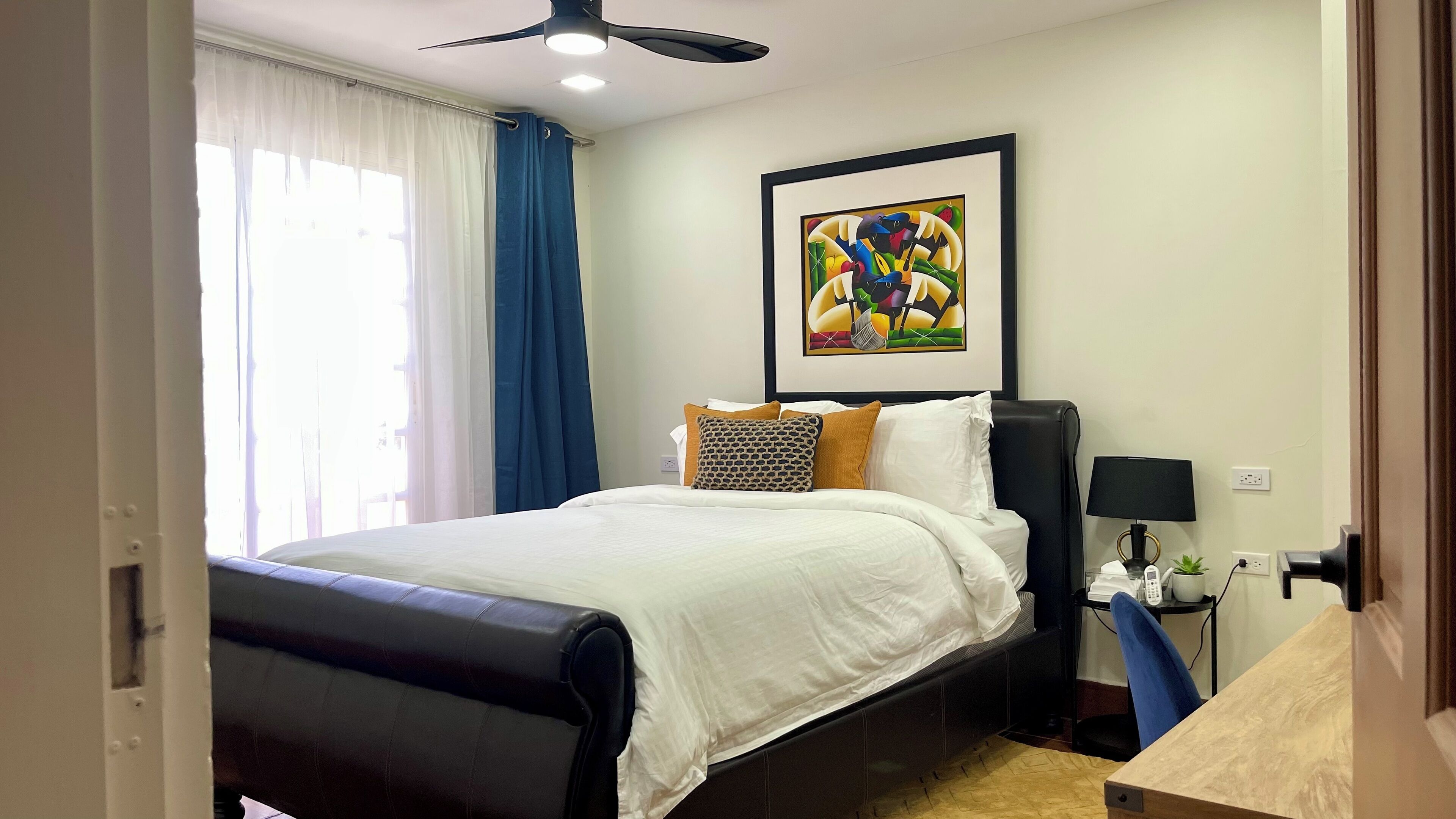 Une chambre d’hôtel avec un lit, un bureau, une chaise, un ventilateur de plafond, un tableau et une fenêtre avec des rideaux.