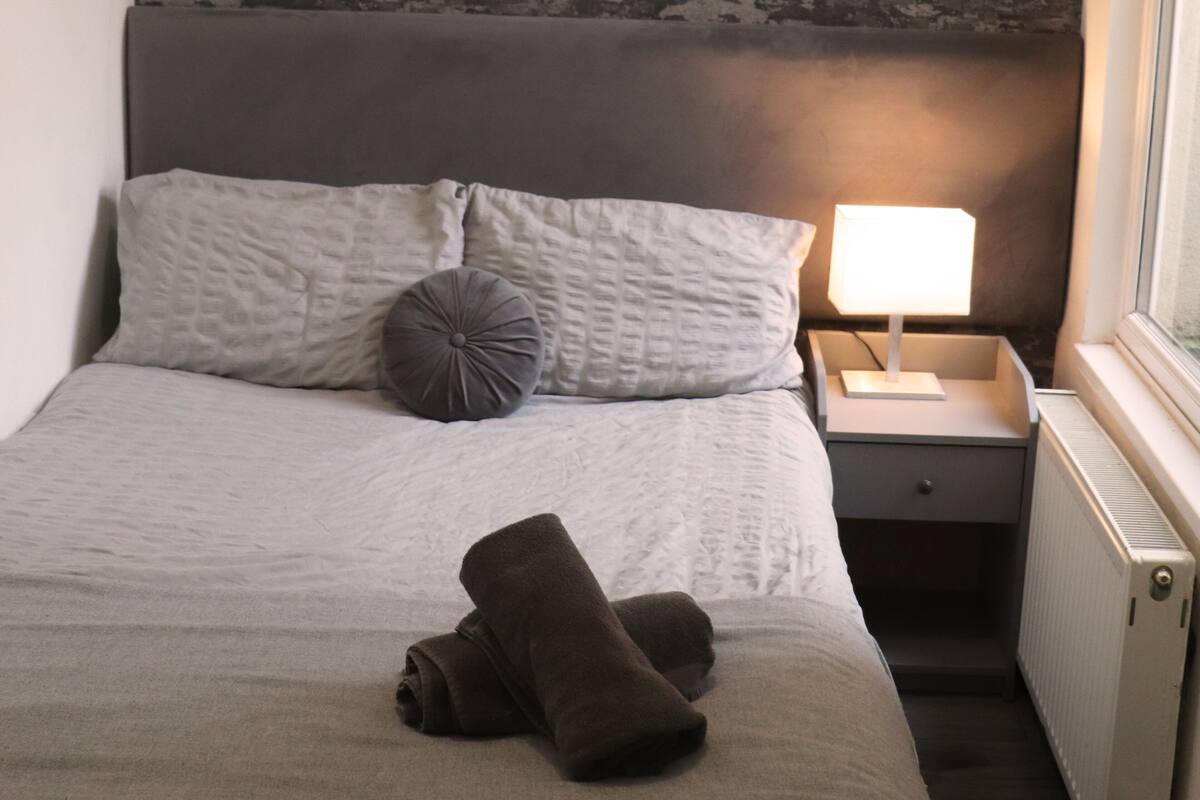 A neatly made bed with pillows and a rolled towel, a bedside lamp, and a radiator.