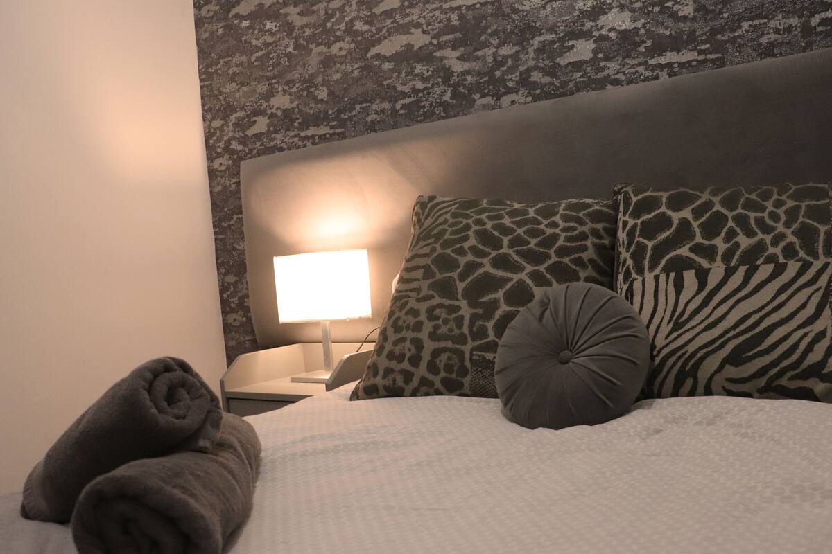 A neatly made bed with a white duvet, two rolled towels, a zebra-patterned pillow, a giraffe-patterned pillow, and a round pillow. A bedside lamp is on to the left.