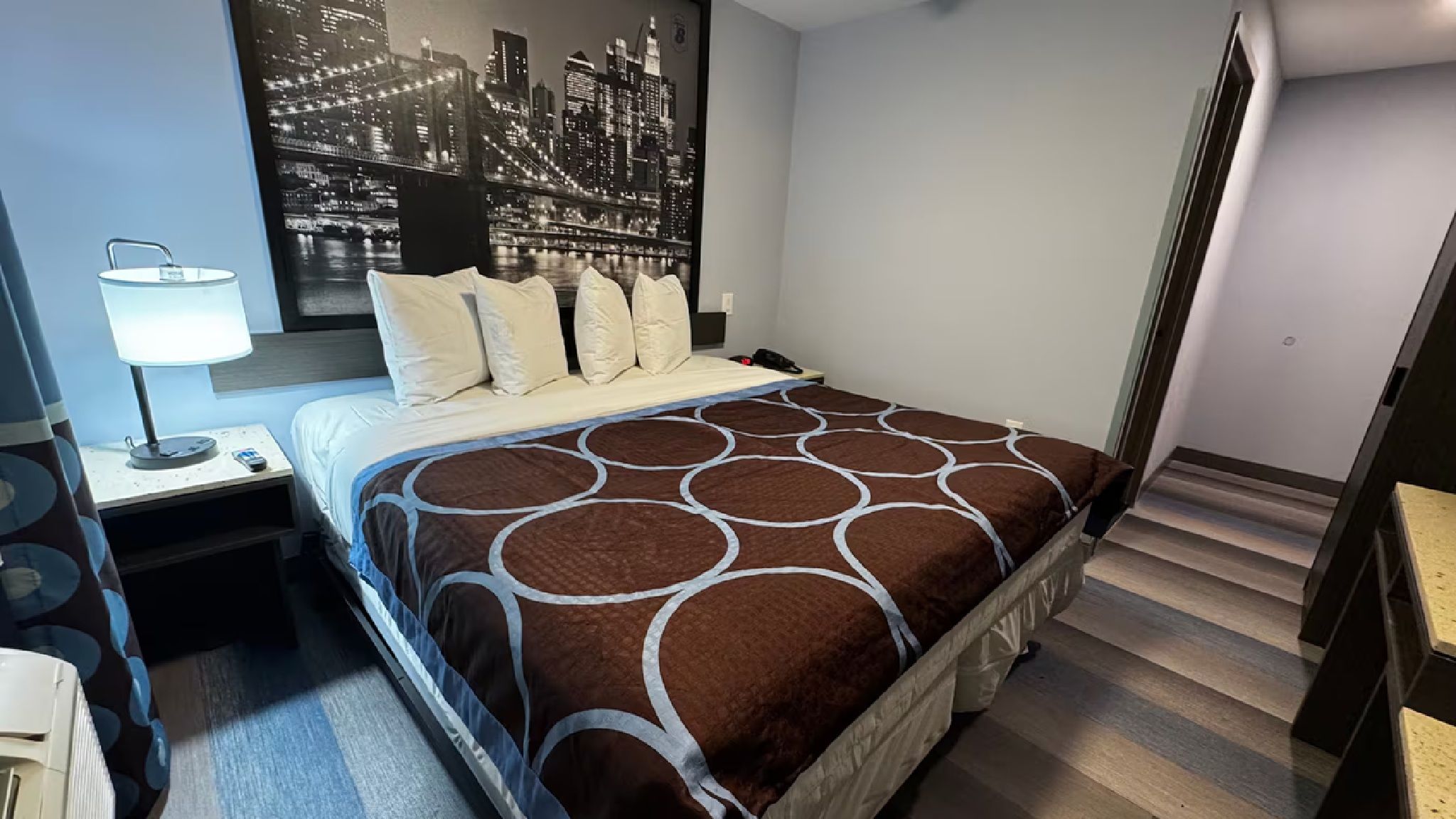 A hotel room with a bed, a nightstand, a lamp, and a cityscape wall mural.