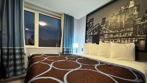 Une chambre d’hôtel avec un grand lit, une fresque représentant un paysage urbain, une fenêtre avec des rideaux et une vue sur le ciel nocturne.