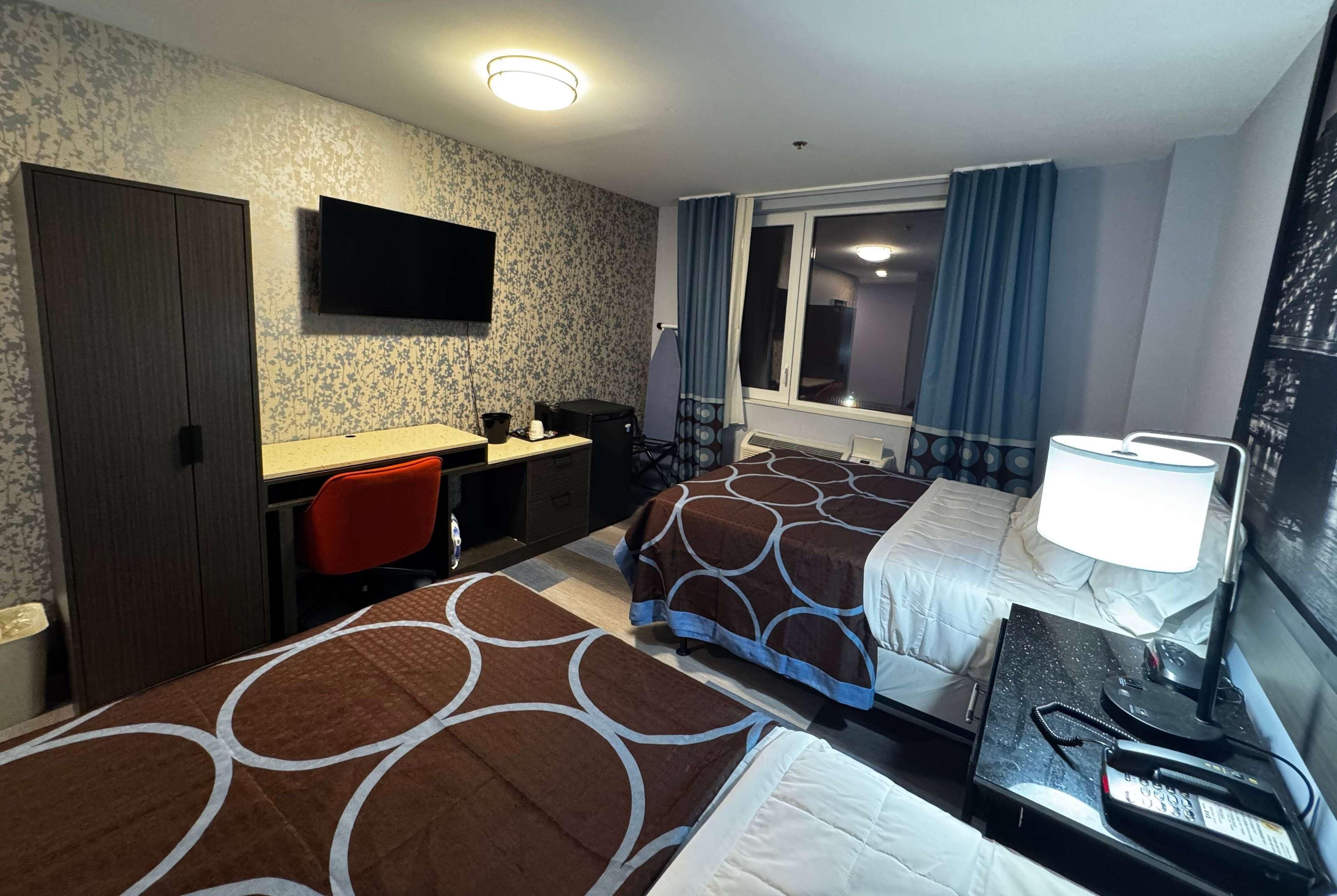 A hotel room with two beds, a desk with a chair, a TV mounted on the wall, a window with curtains, and a nightstand with a lamp and phone.