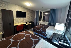 Habitación de hotel con dos camas, un escritorio con silla, televisor montado en la pared, ventana con cortinas y mesita de noche con lámpara y teléfono.