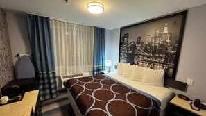 A hotel room with a bed, a desk, a chair, a nightstand, a lamp, and a cityscape view. - Super 8 By Wyndham Brooklyn Fulton Street (Brooklyn)
