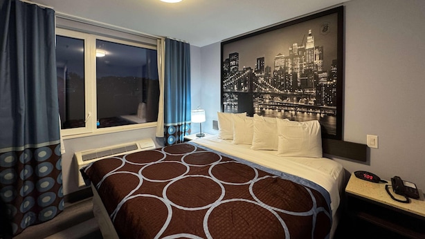A hotel room with a bed, a window with curtains, a cityscape artwork on the wall, a nightstand with a phone, and a wall socket.