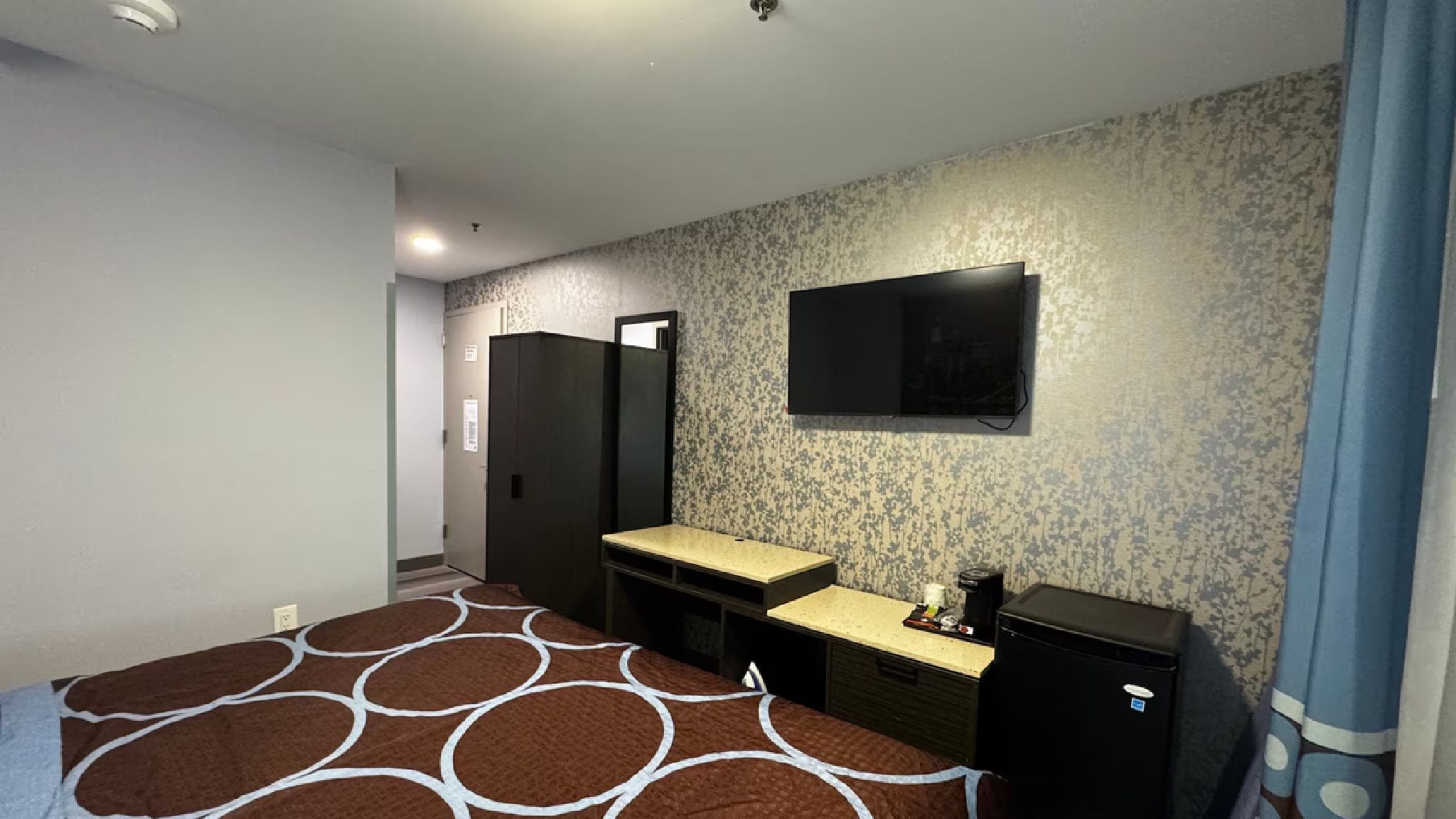 A hotel room with a bed, a desk, a chair, a TV mounted on the wall, and a wardrobe.