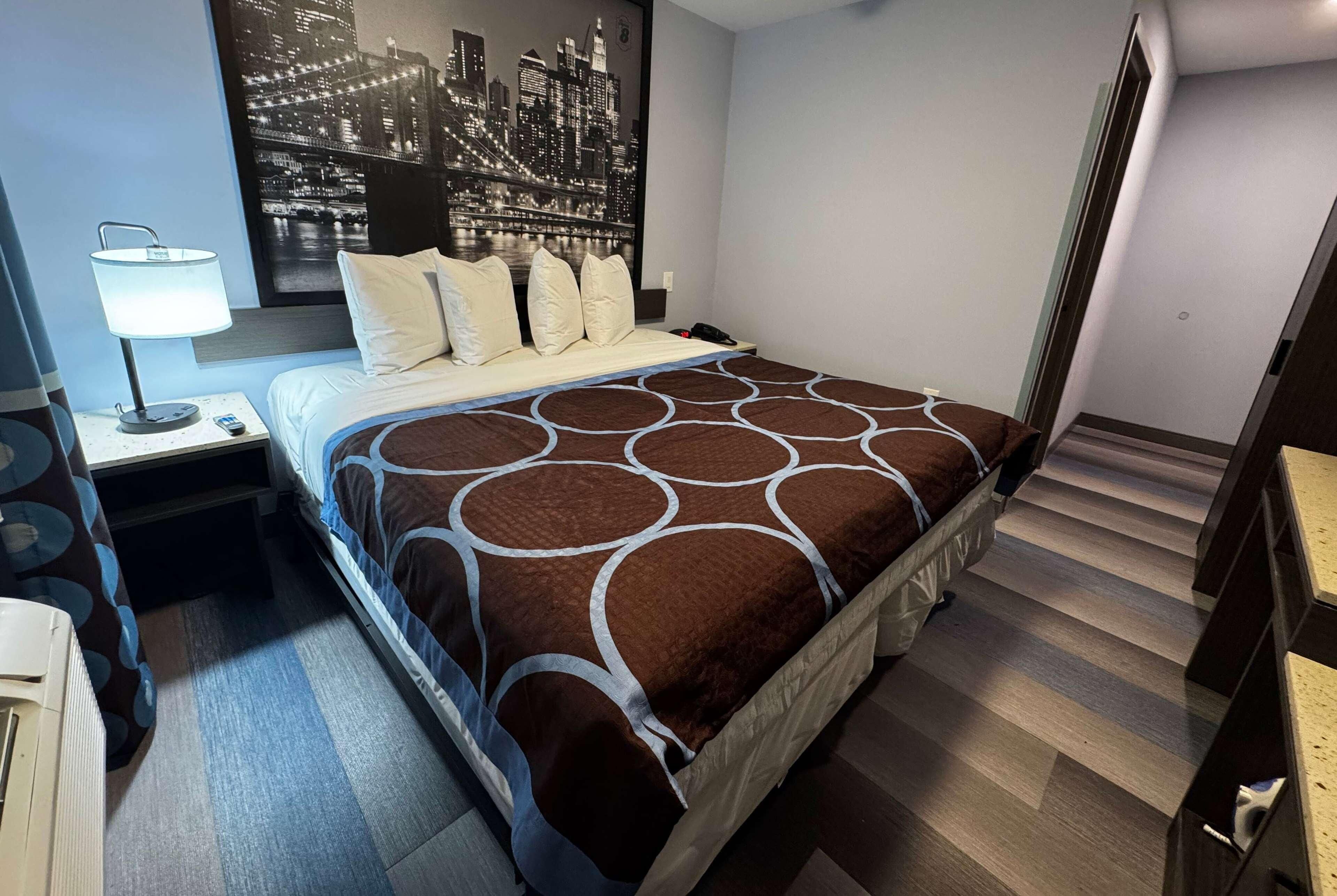 A hotel room with a bed, a nightstand, a lamp, a phone, and a cityscape wall art.