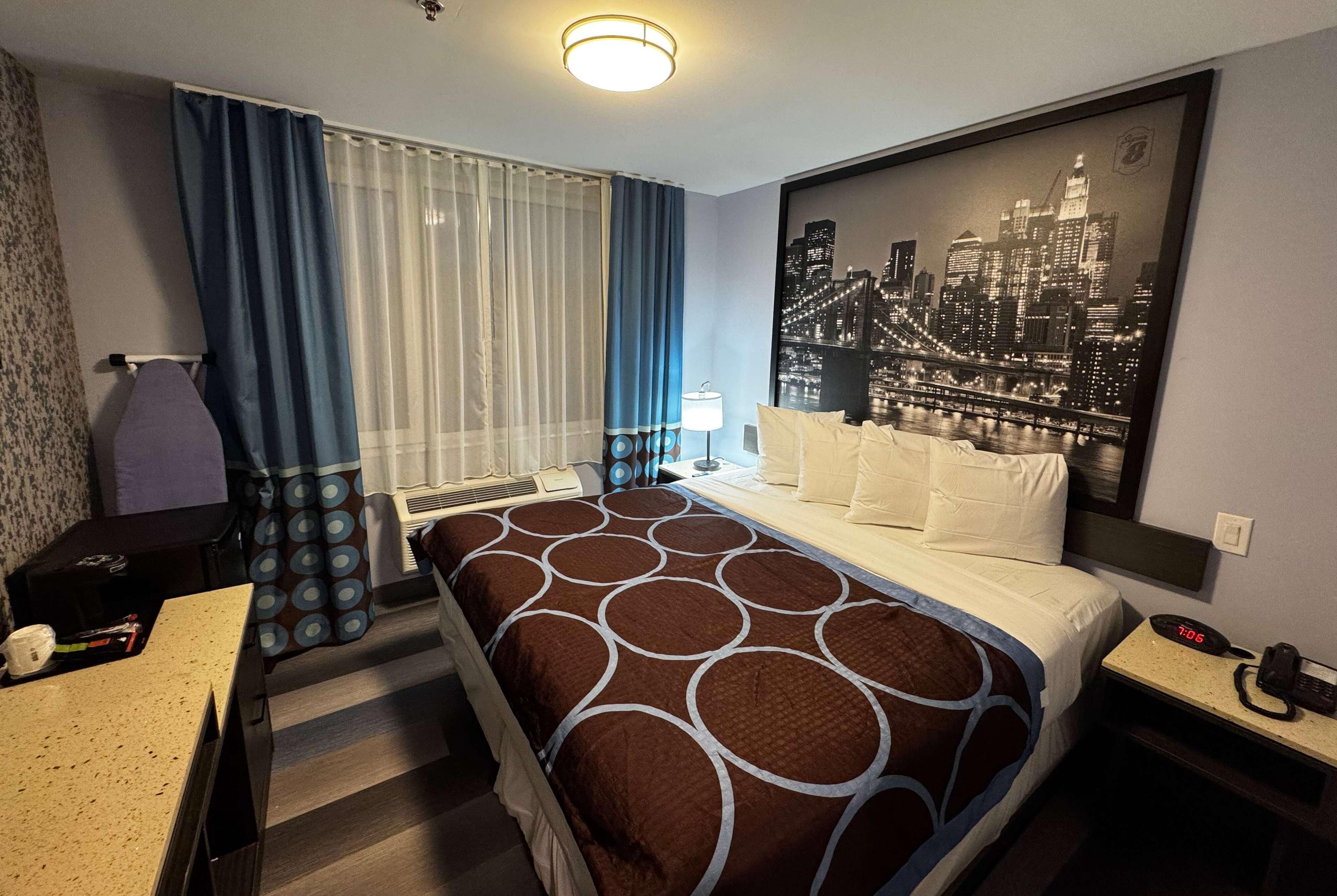A hotel room with a bed, a desk, a chair, a nightstand, a lamp, and a cityscape artwork on the wall.