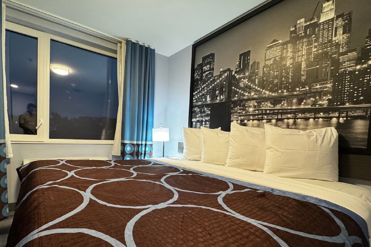 A hotel room with a large bed, a cityscape mural on the wall, and a window with curtains.