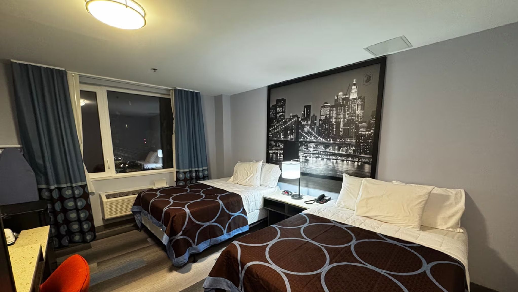 A hotel room with two beds, a desk, a chair, a window with curtains, and a cityscape painting on the wall.