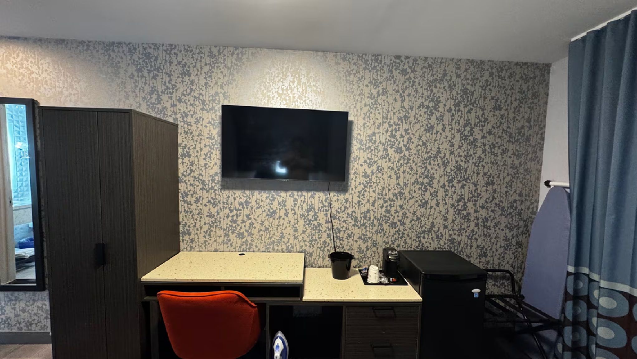 A small room with a TV mounted on the wall, a desk with a chair, a small kitchenette, and a wardrobe.